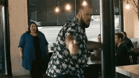 a man and a woman are dancing in a restaurant . the man is wearing a floral shirt .
