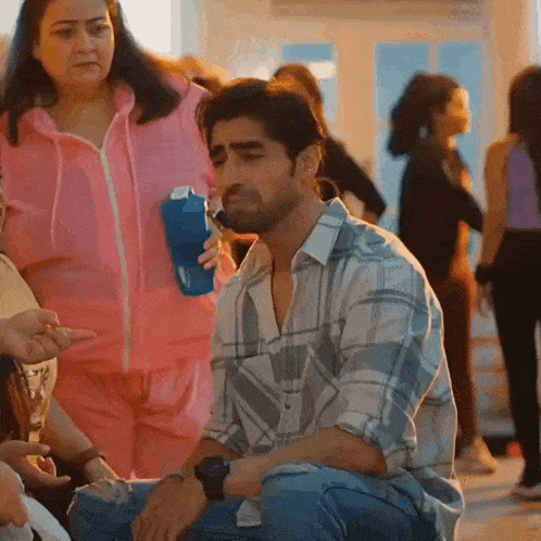a man in a plaid shirt is kneeling down while a woman in a pink jacket holds a blue water bottle