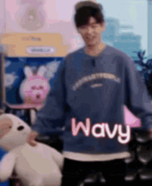 a man wearing a blue sweater is holding a stuffed animal and says wavy .