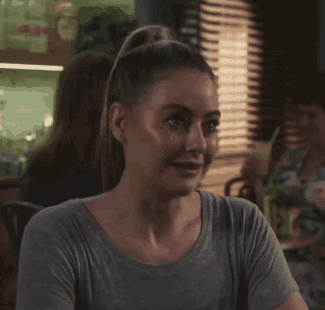 a woman in a gray shirt is smiling in front of a window with blinds