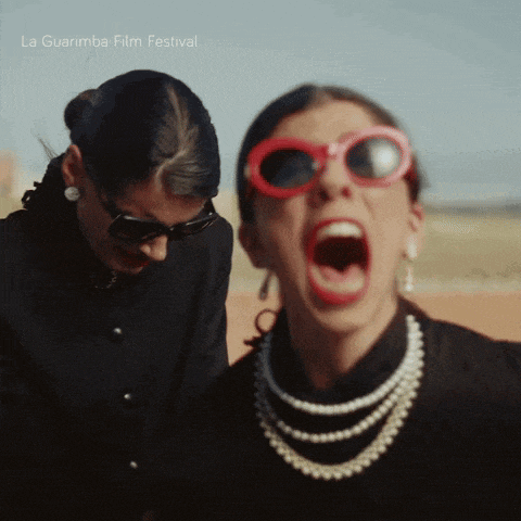 two women wearing pearls and red sunglasses are screaming in front of a la guarimba film festival banner