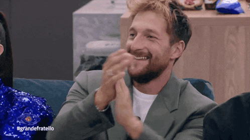 a man sitting on a couch applauding with #grandefratello on the bottom