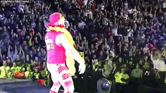 a wrestler in a pink shirt and white pants is walking in front of a crowd .