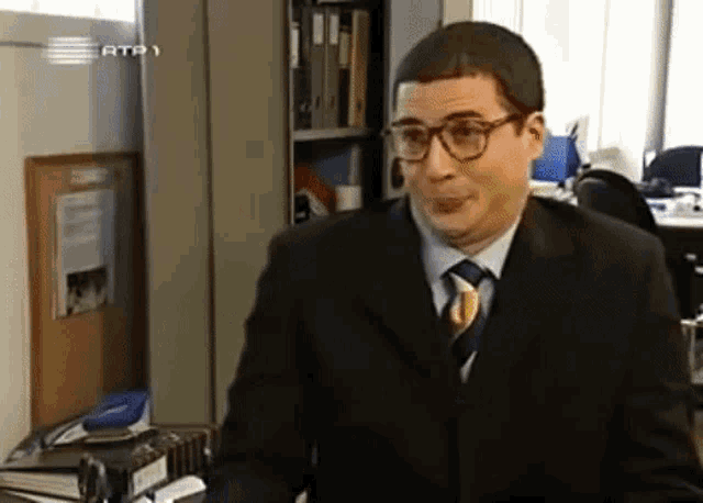 a man in a suit and tie is sitting at a desk in front of a shelf that says rtp 1