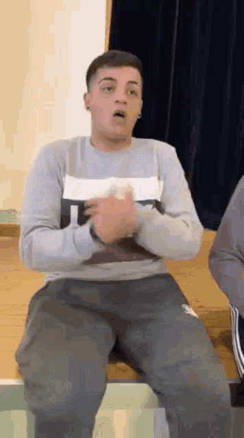 a young man is sitting on a bench with his hands on his chest and making a funny face .
