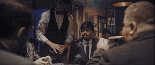 a man in a tuxedo sits at a table while a waiter serves him