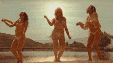 three women in bikinis are dancing on the beach near a body of water