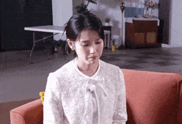 a woman in a white lace shirt is sitting on an orange couch and crying .