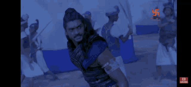 a man in a blue armor is standing in front of a group of people with a subscribe button on the bottom right