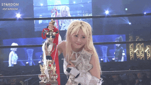 a woman in a wrestling ring holding a trophy with the word stardom on the screen behind her