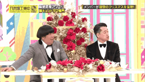two men in tuxedos sit at a table with a christmas tree in the background