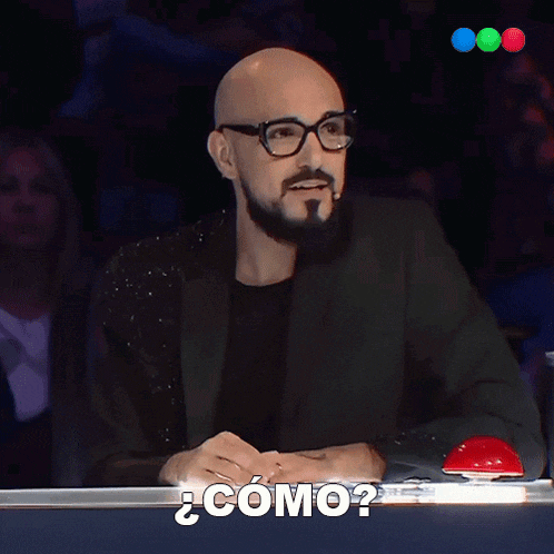 a bald man with glasses and a beard is sitting at a table with a red button that says " como "