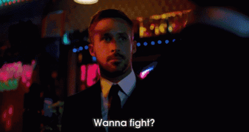 a man in a suit and tie is standing in a dark room and saying wanna fight ?