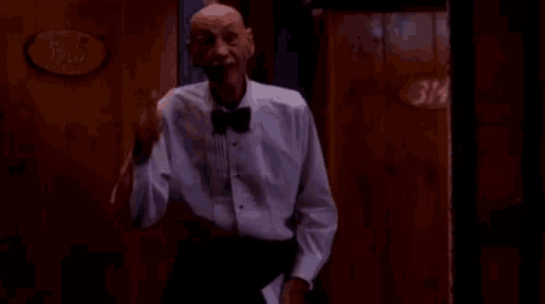 an elderly man in a tuxedo and bow tie is standing in front of a door and giving the middle finger .