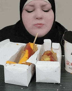 a woman in a hijab is eating a hot dog in a cardboard box