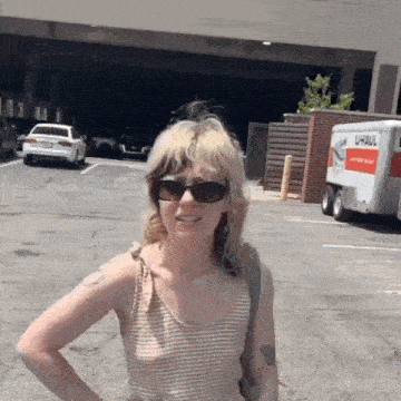 a woman wearing sunglasses and a tank top is standing in a parking lot with her hands on her hips .