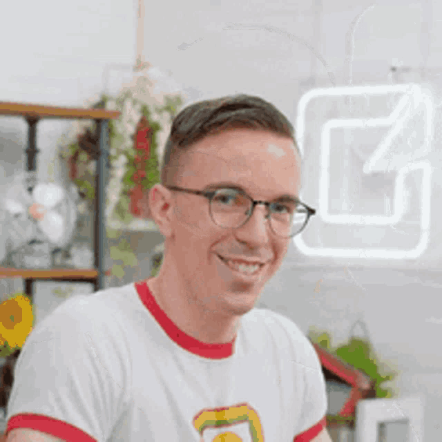 a man wearing glasses and a white shirt is smiling while sitting in front of a neon sign .