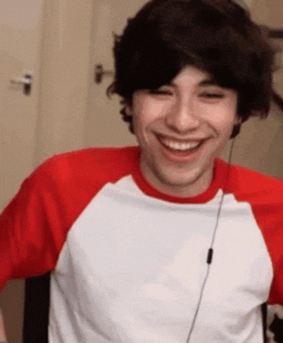 a young man wearing a red and white t-shirt is smiling while sitting in a chair .