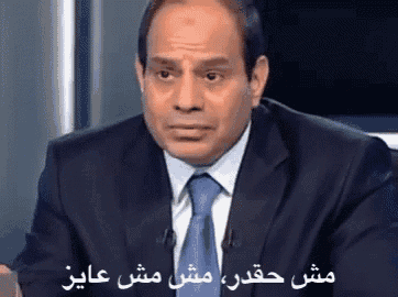 a man in a suit and tie is sitting in front of a microphone with arabic writing behind him .