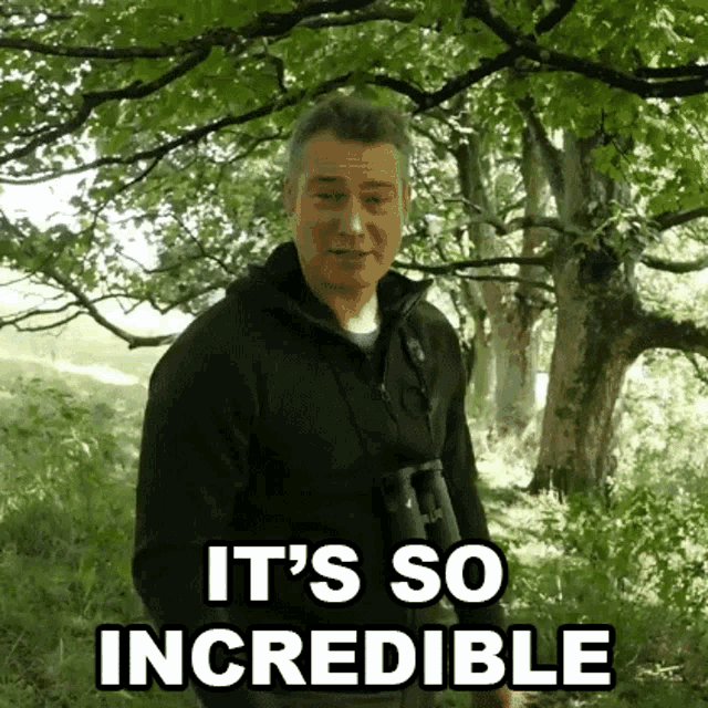 a man in a black jacket is standing in the woods holding binoculars and says it 's so incredible .