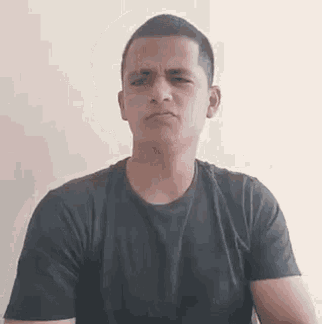 a man in a black t-shirt is making a funny face while sitting in front of a wall .