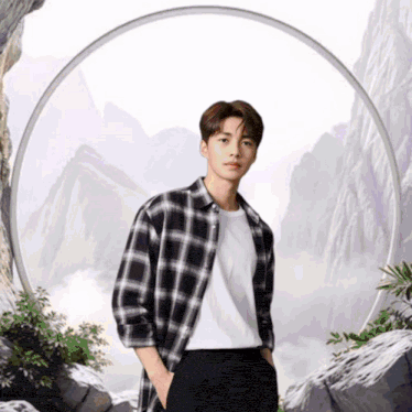 a young man wearing a plaid shirt and a white shirt stands in front of a mountain