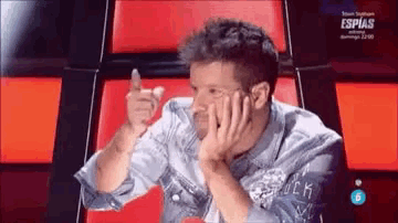 a man in a denim shirt is sitting in a red chair with his hand on his face