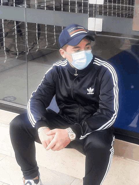 a man wearing a face mask and an adidas jacket sits on the sidewalk