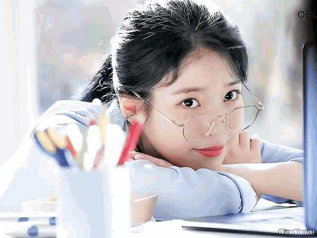 a woman wearing glasses sits at a desk with her head resting on her elbow