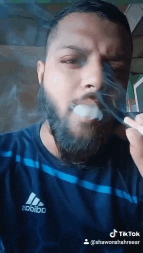 a man with a beard wearing an adidas shirt is smoking a cigarette