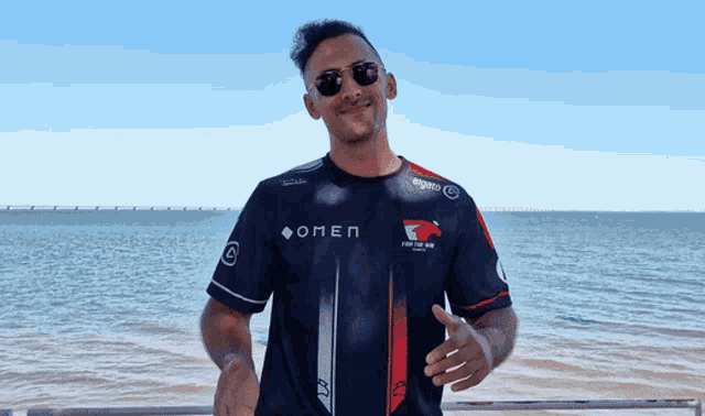 a man wearing sunglasses and a shirt that says omen stands in front of the ocean