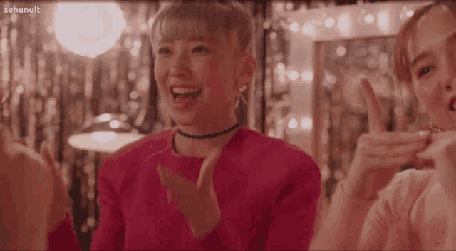 a woman in a pink sweater is clapping her hands in front of a sign that says " sehunult "