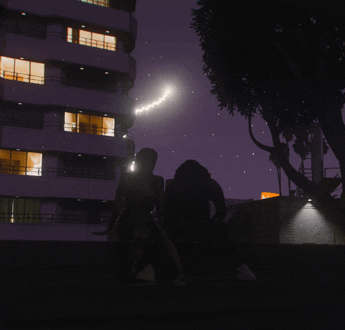 a man and a woman sit in front of a building at night with a tree in the foreground
