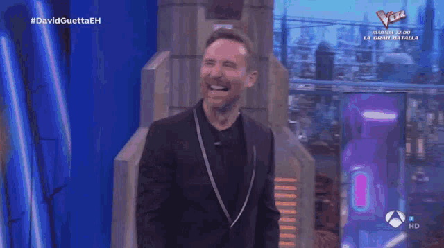 a man in a black suit is laughing in front of a sign that says #davidguettaeh