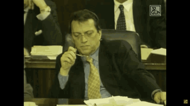 a man in a suit and tie sits at a desk with the number 32 gun on the bottom right