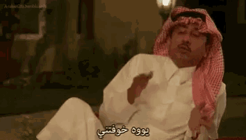 a man in a keffiyeh is sitting on a couch with his eyes closed and making a funny face .