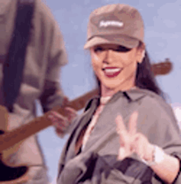 a woman wearing a supreme hat is giving a peace sign while a man plays a guitar .