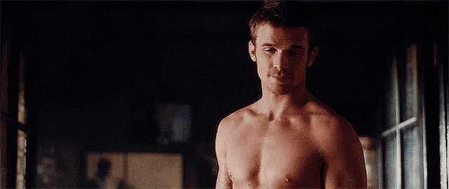 a shirtless man is standing in a dark room next to a window .