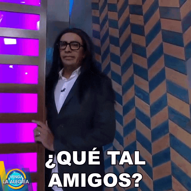 a man with long hair and glasses stands in front of a wall and says " que tal amigos "