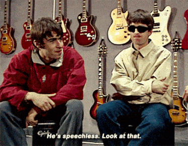 two men are sitting in front of a wall of guitars with one saying he 's speechless look at that