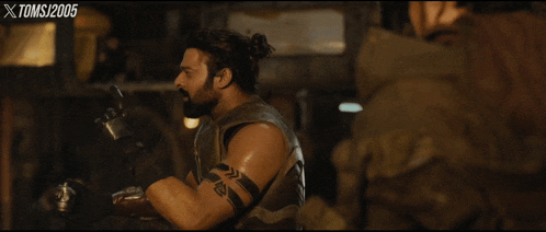 a man with a ponytail and a tattoo on his arm is in a dark room with xtoms written on the bottom
