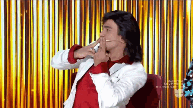 a man in a white jacket and red shirt is sitting in front of a gold curtain .