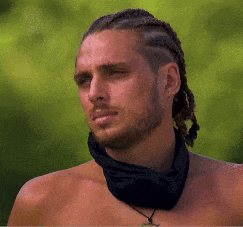 a shirtless man with braided hair and a bandana around his neck looks at the camera