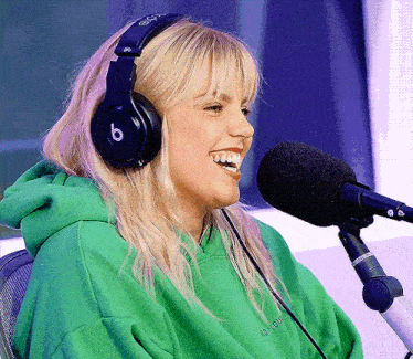 a woman wearing headphones and a green hoodie is smiling in front of a microphone