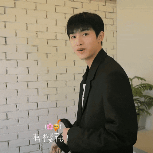 a man in a black jacket smells something in front of a white brick wall with chinese writing on it