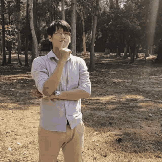 a man in a purple shirt and tan pants stands in a park with his arms crossed