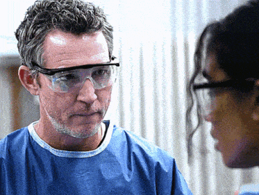 a man wearing goggles talks to a woman in a lab coat