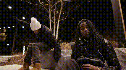 two men are sitting next to each other on a bench at night . one of the men is wearing a white hat .
