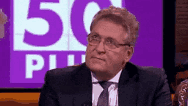 a man in a suit and tie is sitting in front of a purple 50 plus sign