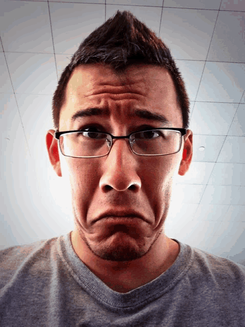 a man wearing glasses and a grey shirt is making a sad face
